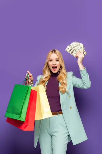 Happy blonde young woman in fashionable turquoise blazer with shopping bags and money on purple background — Stock Photo