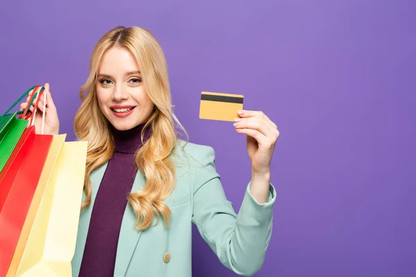 Sonriente joven rubia en blazer turquesa de moda con bolsas de compras y tarjeta de crédito sobre fondo púrpura - foto de stock