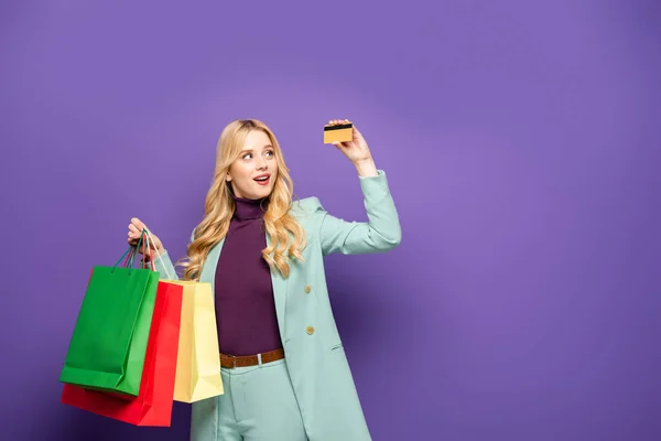 Mujer joven rubia en blazer turquesa de moda con bolsas de compras y tarjeta de crédito sobre fondo púrpura - foto de stock