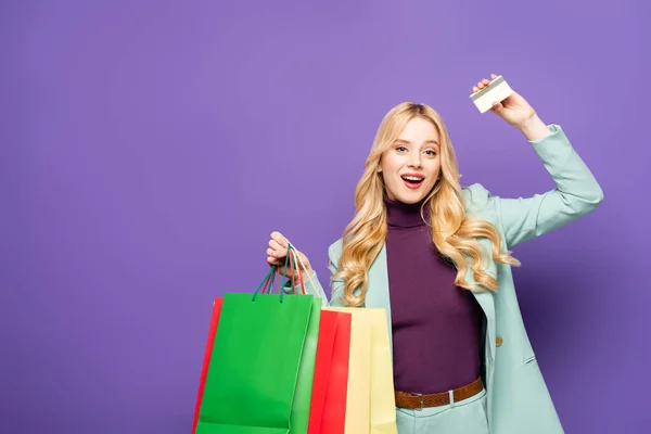 Happy blonde young woman in fashionable turquoise blazer with shopping bags and credit card on purple background — Stock Photo