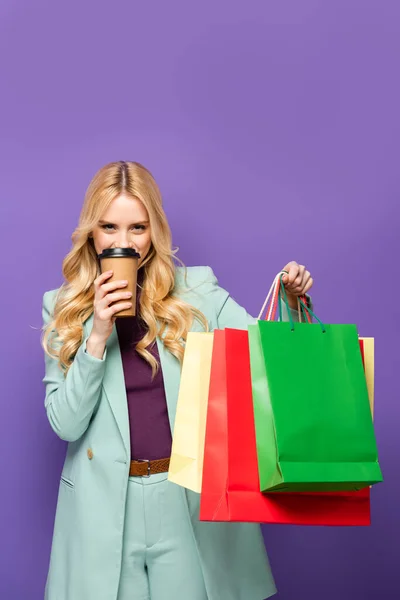 Mujer joven rubia en blazer turquesa de moda con bolsas de compras y taza de papel sobre fondo morado - foto de stock