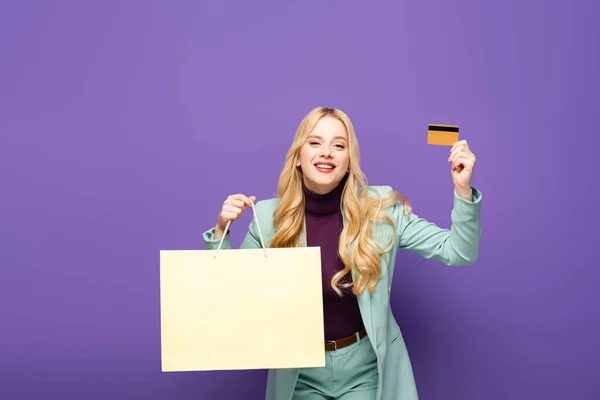 Happy blonde young woman in fashionable turquoise blazer with credit card and shopping bag on purple background — Stock Photo