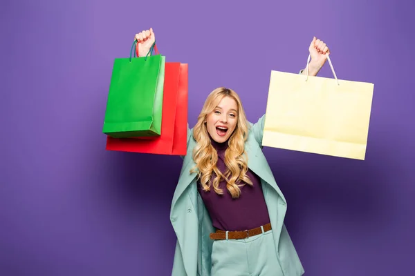 Glückliche blonde junge Frau im modischen türkisfarbenen Blazer mit Einkaufstaschen auf lila Hintergrund — Stockfoto