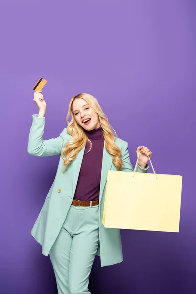Glückliche blonde junge Frau im modischen türkisfarbenen Blazer mit Kreditkarte und Einkaufstasche auf lila Hintergrund — Stockfoto
