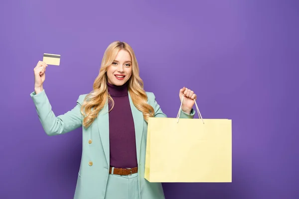 Happy blonde young woman in fashionable turquoise blazer with credit card and shopping bag on purple background — Stock Photo