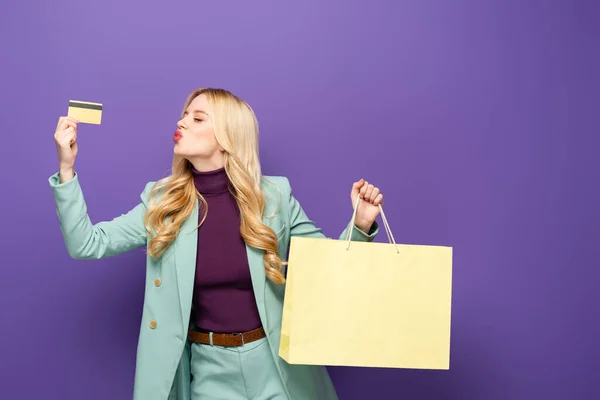 Blonde young woman in fashionable turquoise blazer with credit card and shopping bag showing kiss on purple background — Stock Photo