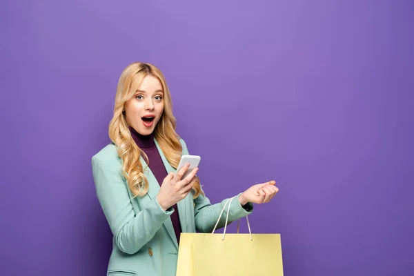Shocked blonde young woman in fashionable turquoise blazer with smartphone and shopping bag on purple background — Stock Photo