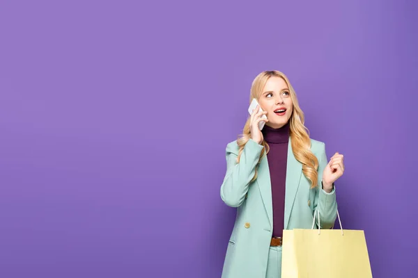 Blonde young woman in fashionable turquoise blazer talking on smartphone and holding shopping bag on purple background — Stock Photo