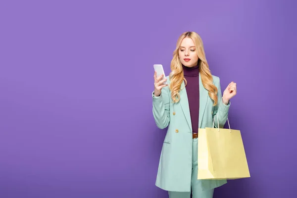 Jovem loira em blazer turquesa elegante com smartphone e saco de compras no fundo roxo — Fotografia de Stock