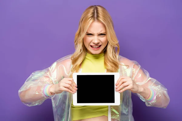 Mujer joven rubia confundida en traje colorido presentando tableta digital sobre fondo púrpura - foto de stock