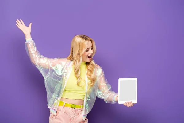 Mujer joven rubia feliz en traje colorido presentando tableta digital sobre fondo púrpura - foto de stock