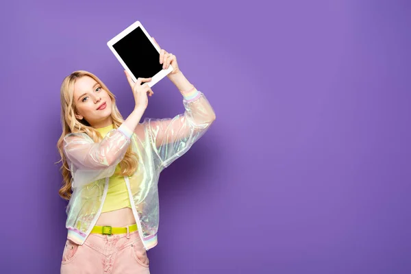 Mujer joven rubia en traje colorido presentando tableta digital sobre fondo púrpura - foto de stock