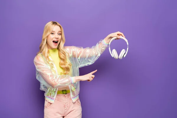 Excited blonde young woman in colorful outfit pointing at headphones on purple background — Stock Photo