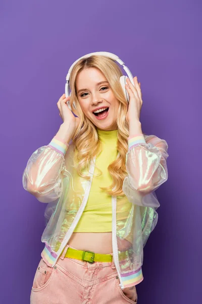 Happy blonde young woman in colorful outfit and headphones on purple background — Stock Photo