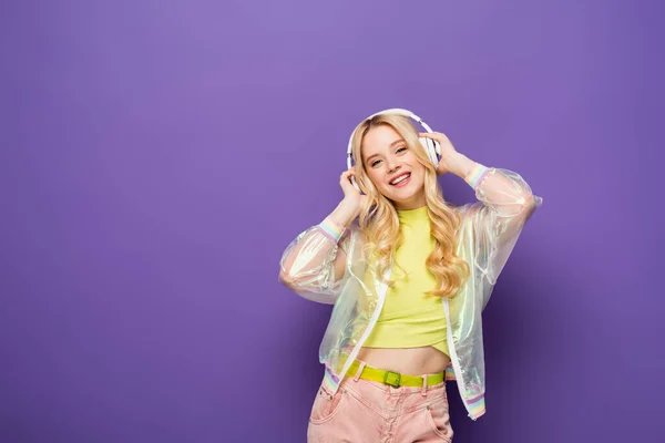 Mujer joven rubia feliz en traje colorido y auriculares sobre fondo morado - foto de stock