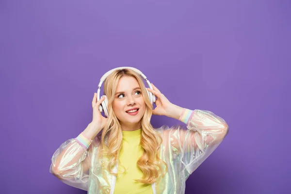 Glückliche blonde junge Frau in buntem Outfit und Kopfhörer auf lila Hintergrund — Stockfoto