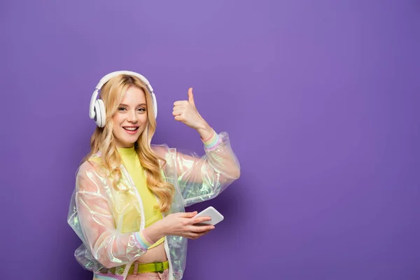 Happy blonde young woman in colorful outfit and headphones holding smartphone and showing thumb up on purple background — Stock Photo