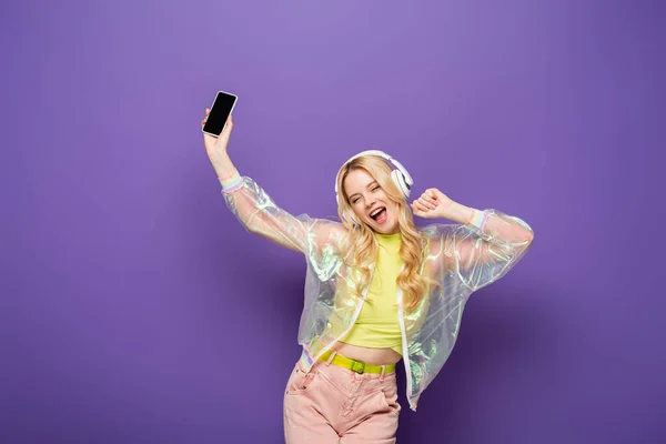 Happy blonde young woman in colorful outfit and headphones holding smartphone on purple background — Stock Photo