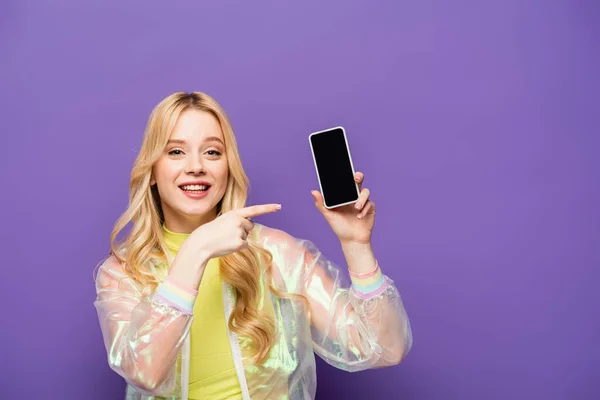 Smiling blonde young woman in colorful outfit pointing at smartphone on purple background — Stock Photo