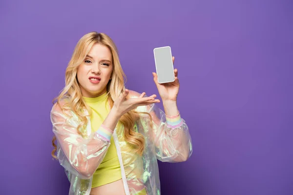 Смущенная блондинка в красочном наряде представляет смартфон на фиолетовом фоне — стоковое фото