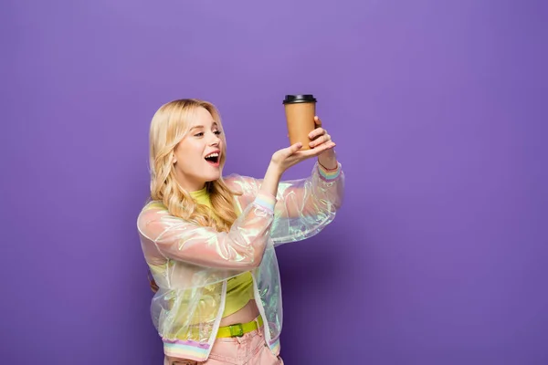 Aufgeregte blonde junge Frau in buntem Outfit mit Pappbecher auf lila Hintergrund — Stockfoto