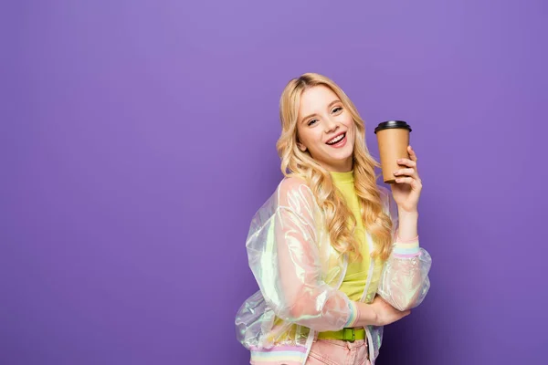 Happy blonde young woman in colorful outfit with paper cup on purple background — Stock Photo