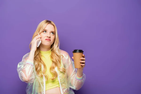 Traurige blonde junge Frau in buntem Outfit mit Pappbecher im Gespräch auf Smartphone auf lila Hintergrund — Stockfoto