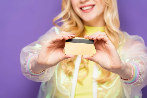 Cropped view of happy blonde young woman in colorful outfit showing credit card on purple background — Stock Photo