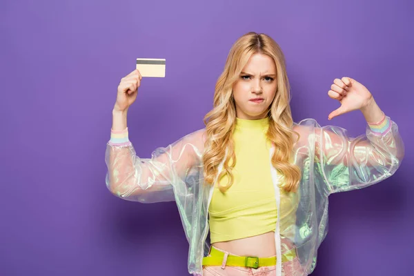 Сумна блондинка молода жінка в барвистому вбранні, що показує кредитну картку і великий палець вниз на фіолетовому фоні — стокове фото