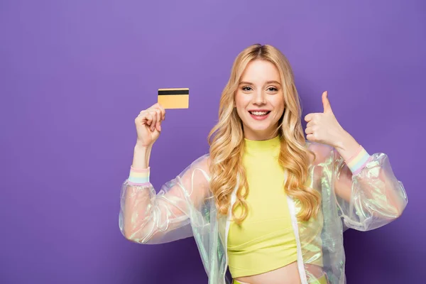 Happy blonde young woman in colorful outfit showing credit card and thumb up on purple background — Stock Photo