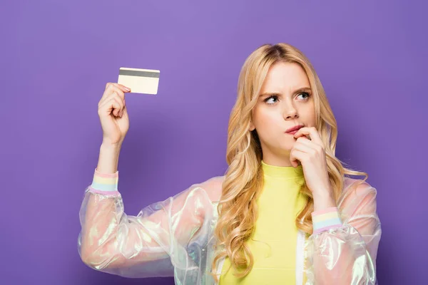 Mujer joven rubia pensativa en traje colorido que muestra la tarjeta de crédito sobre fondo púrpura - foto de stock