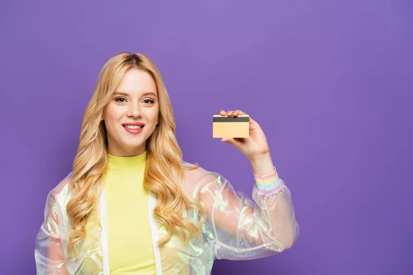 Sorrindo loira jovem em roupa colorida mostrando cartão de crédito no fundo roxo — Fotografia de Stock
