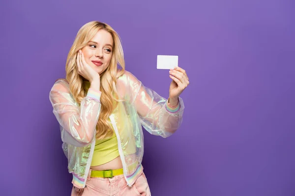 Bonita loira jovem em roupa colorida segurando cartão em branco no fundo roxo — Fotografia de Stock