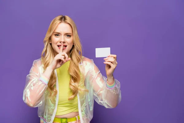 Jovem loira em roupa colorida segurando cartão em branco e mostrando sinal de shh no fundo roxo — Fotografia de Stock