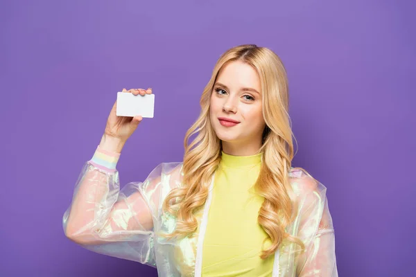 Smiling blonde young woman in colorful outfit holding blank card on purple background — Stock Photo