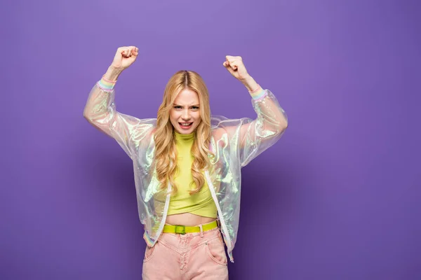 Bionda giovane donna in abito colorato con pugni alzati su sfondo viola — Foto stock