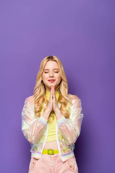 Blonde young woman in colorful outfit with praying hands on purple background — Stock Photo