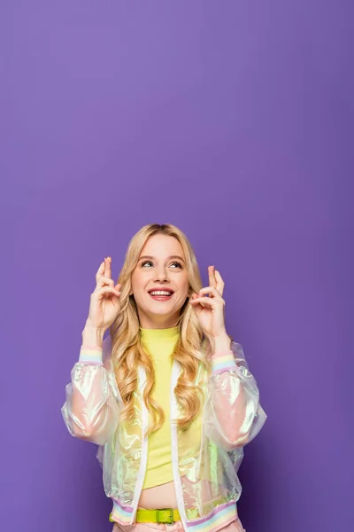 Happy blonde young woman in colorful outfit with crossed fingers on purple background — Stock Photo