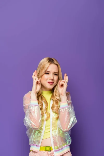 Blonde young woman in colorful outfit with crossed fingers on purple background — Stock Photo