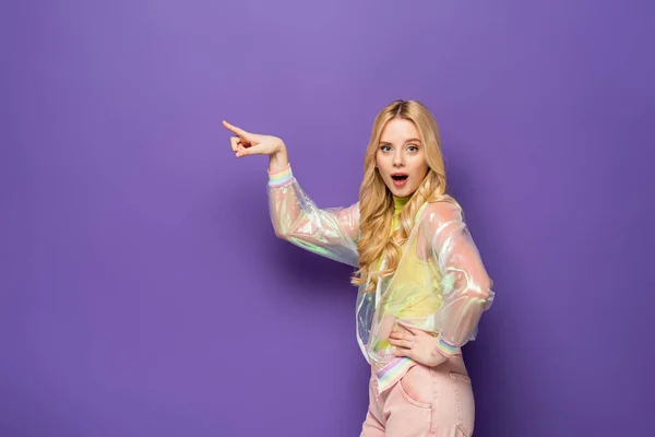 Side view of shocked blonde young woman in colorful outfit pointing aside on purple background — Stock Photo