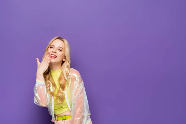Jeune femme blonde souriante en tenue colorée avec la main près du visage sur fond violet — Photo de stock