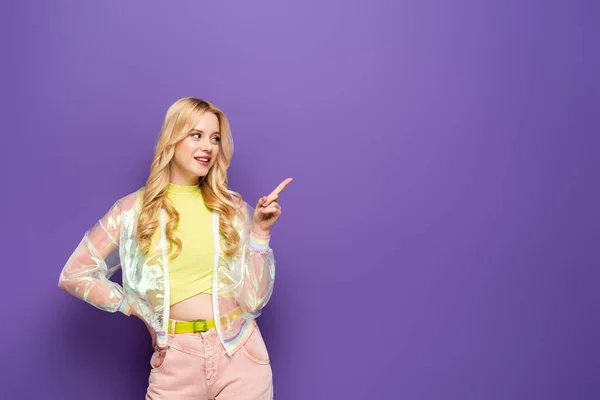 Souriant blonde jeune femme en tenue colorée pointant de côté sur fond violet — Photo de stock