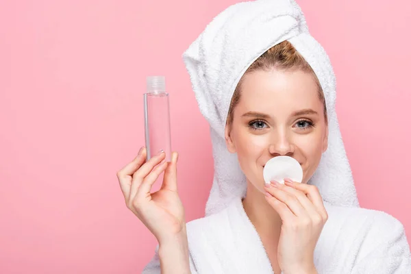 Jovem mulher em roupão de banho com toalha na cabeça com água micelar e algodão pad isolado em rosa — Fotografia de Stock