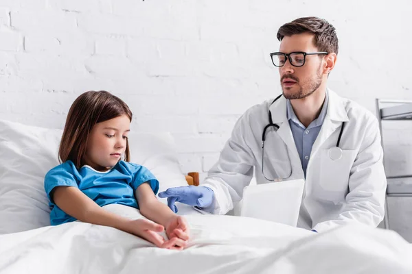 Pediatrician with digital tablet talking to child sitting on bed in hospital — Stock Photo