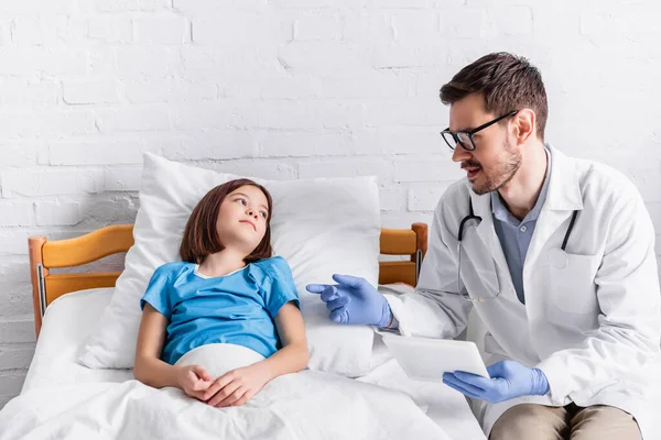 Pediatra con tableta digital hablando con una chica sonriente acostada en la cama en la clínica - foto de stock