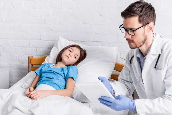 Medico che indica tablet digitale mentre parla con bambino malato sdraiato a letto — Foto stock