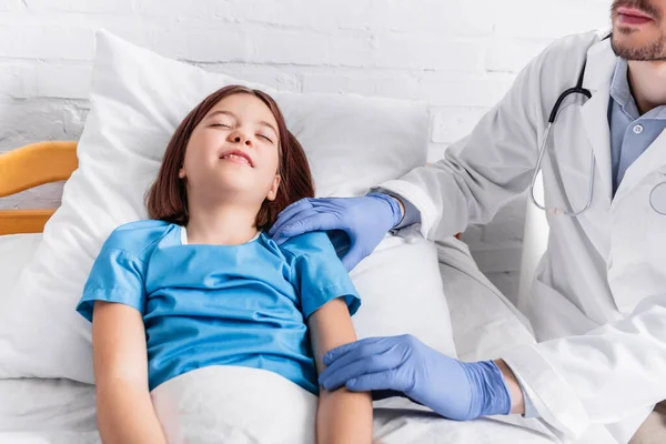 Pediatrician examining diseased girl lying in bed with closed eyes — Stock Photo