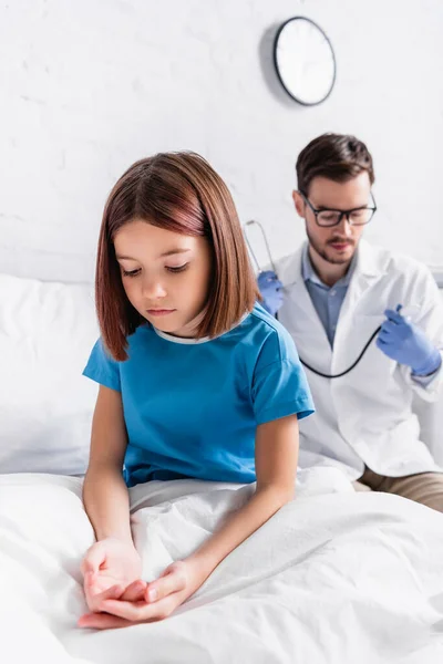 Diseased girl sitting on bed in clinic near pediatrician with stethoscope on blurred background — Stock Photo