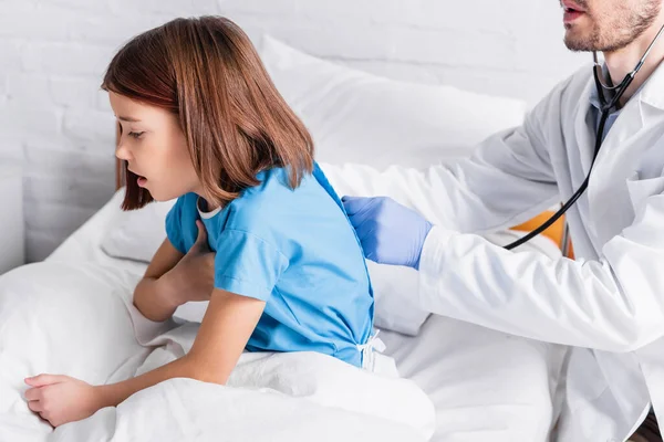 Krankes Mädchen hustet im Bett, während der Arzt sie mit Stethoskop untersucht — Stockfoto