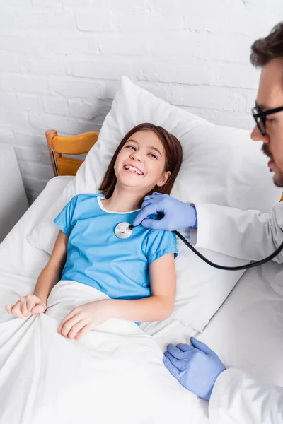 Kinderarzt untersucht fröhliches Mädchen mit Stethoskop im Krankenhaus, unscharfer Vordergrund — Stockfoto